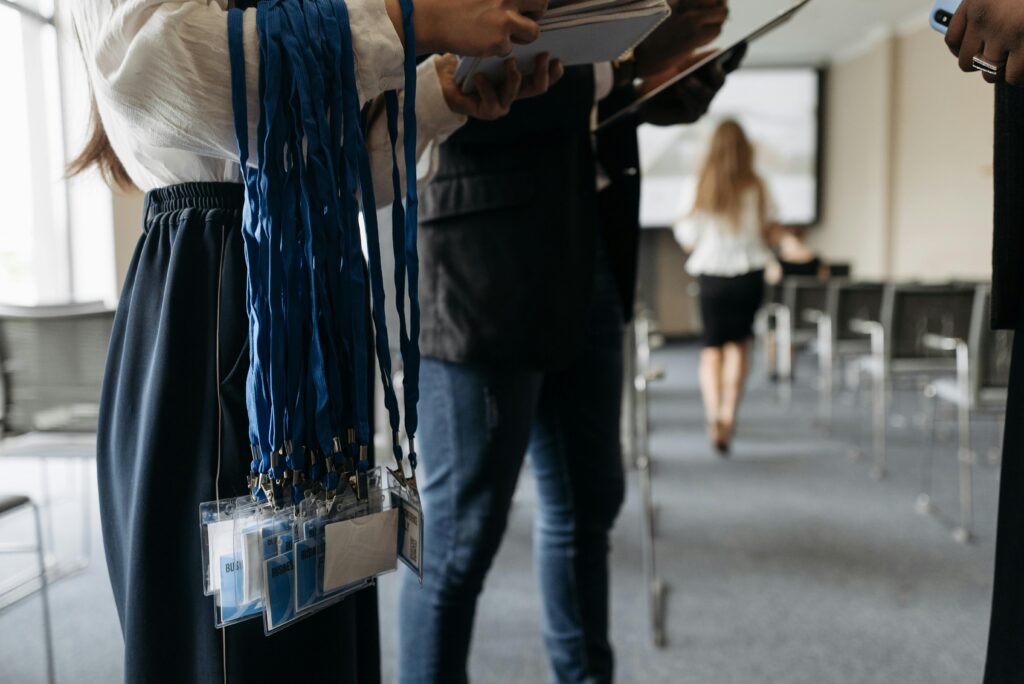 Badge: Usi, tipologie e benefici. Nell'immagine due persone si accingono a distribuire i badge, che pendono dal braccio destro di una delle due, ai partecipanti ad un convegno, all'ingresso dell'aula preposta ad accoglierlo.